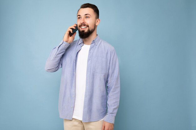 Fotoaufnahme eines glücklichen, gut aussehenden jungen bärtigen Mannes, der ein lässiges blaues Hemd und ein weißes T-Shirt trägt