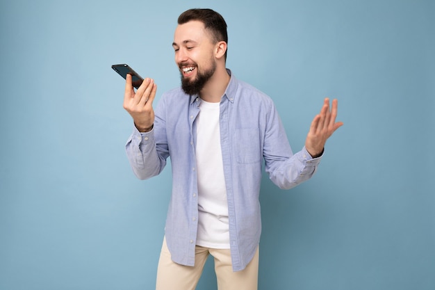 Fotoaufnahme eines gutaussehenden, positiv aussehenden jungen Mannes in einem lässigen, stilvollen Outfit, das isoliert auf dem Hintergrund mit leerem Raum in der Hand steht und das Mobiltelefon verwendet, um Sprachnachrichten aufzunehmen