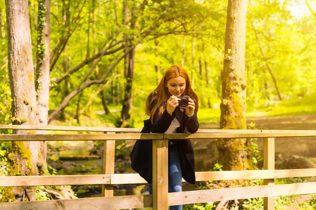 Fotógrafa com uma jaqueta preta curtindo em um parque de outono, tirando fotos em uma ponte de madeira