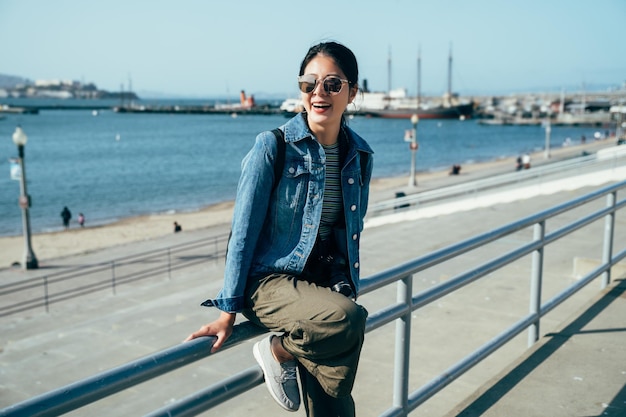 una fotógrafa feliz con gafas de sol disfruta de la hermosa vista del océano azul y el cielo apoyándose en un pasamanos cerca del puerto. joven asiática sonriendo riendo haciendo turismo en el muelle 39.