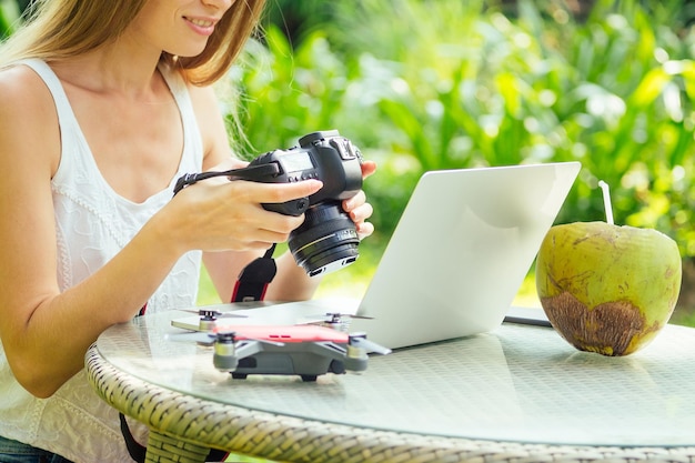 Fotógrafa insertando o quitando una tarjeta de memoria en su cámara dslr profesional mientras se sienta en un escritorio con su computadora portátil en el fondo de palmeras verdes de Paradise Beach