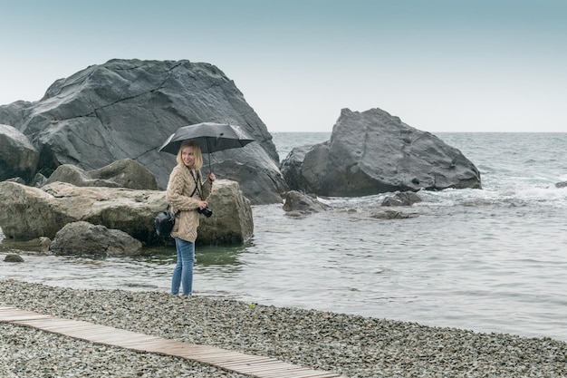 Una fotógrafa con un paraguas de la lluvia hace una foto en la costa pedregosa del mar el océano Fotografiando en todas las condiciones climáticas