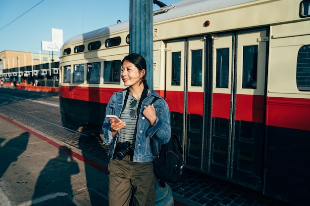 la fotógrafa de viajes se baja del tren ligero sonriendo caminando de regreso al hotel con la puesta de sol. ajetreado concepto de estilo de vida de la ciudad urbana de san francisco. joven asiática sosteniendo un celular chateando en línea.
