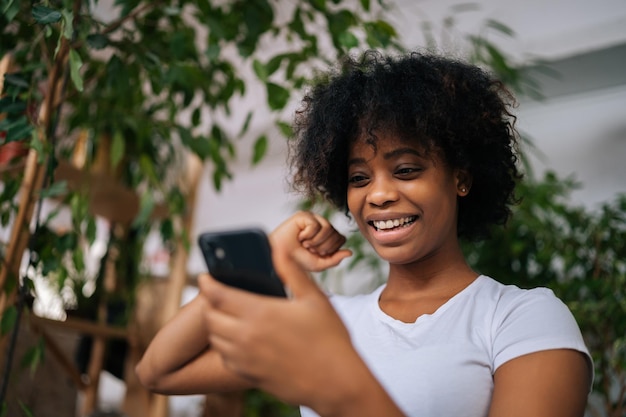 Fotografando por baixo do rosto de uma jovem afro-americana alegre e encaracolada, segurando o celular nas mãos, desfrutando da navegação na web, comunicando-se na rede social