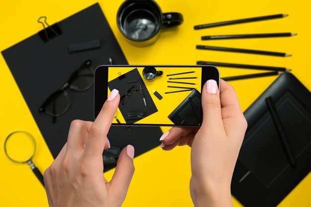 Fotografar artigos de papelaria pretos na câmera do telefone. Artigos de papelaria em fundo amarelo. Mão de uma mulher de close-up segurando o telefone.