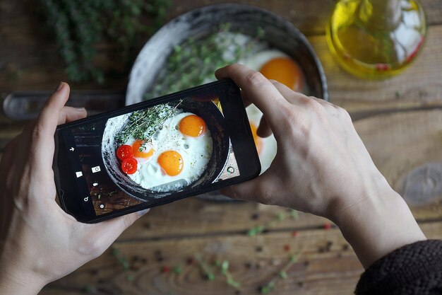 Fotografar comida na câmera do telefone, ovos fritos em uma panela velha com tomates em uma mesa de madeira, foco seletivo, fotógrafo de alimentos, close-up mão segurando o telefone no café da manhã