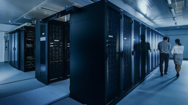 Fotografía de un administrador de TI con una computadora portátil y un joven colega técnico caminando junto a los racks de servidores en el centro de datos ejecutando diagnósticos o mantenimiento