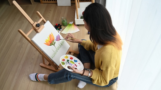 Fotografía aérea de una joven artista pintando sobre lienzo mientras está sentada en el suelo de su estudio.