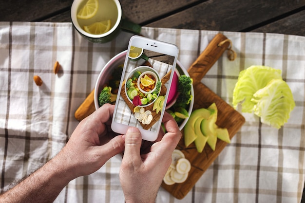 Fotografía de alimentos por teléfono. Concepto de contenido de blogs de alimentos. Manos masculinas toman foto de plato vegano con pantalla de teléfono vista superior