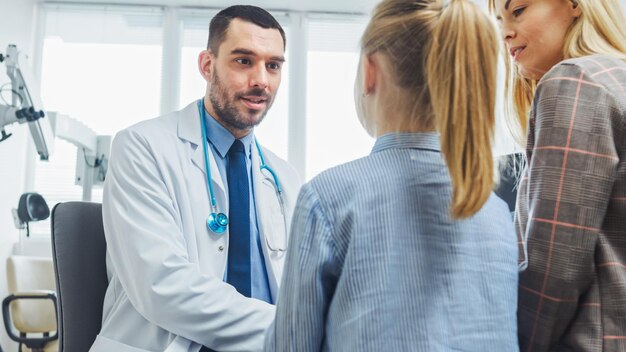 Foto fotografía en bajo ángulo de un amigable médico explicando cómo funciona la medicina a una linda niña y su madre pediatra cura a la niña oficina médica moderna