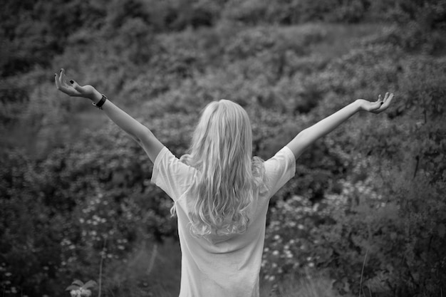 Fotografía en blanco y negro. Atractiva mujer joven rubia con las manos levantadas en el bosque de pie de espaldas a la cámara.