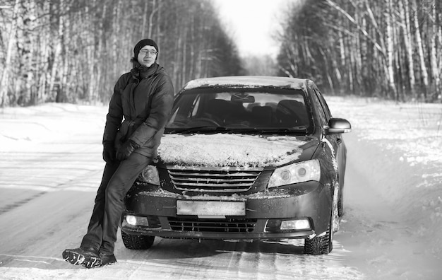 Fotografía en blanco y negro de la carretera rural de invierno en el bosque en un día soleado y el hombre con el coche