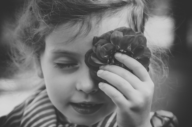 Fotografía en blanco y negro de la niña que cubre su ojo con rojo georgina o dahlia. Hermosa jovencita de piel pura y sonrisa jugando con flor y tocándose la cara con ella.