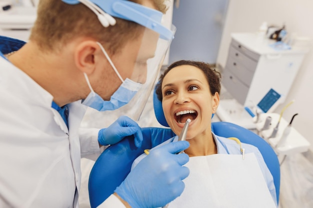 Fotografía cenital del dentista masculino con máscara protectora que examina los dientes del cliente con herramientas dentales en la clínica