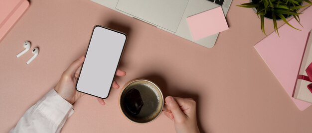 Fotografía cenital de mujer con smartphone con trazado de recorte y sosteniendo la taza de café en la mesa rosa