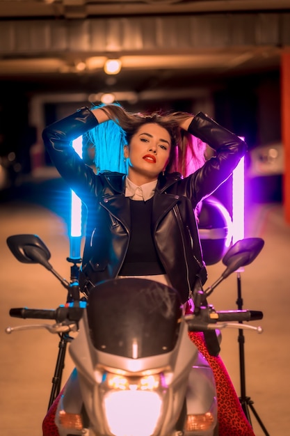 Fotografia com neons azuis e rosa em uma motocicleta. Retrato de uma jovem loira e bonita, branca, com uma jaqueta de couro preta