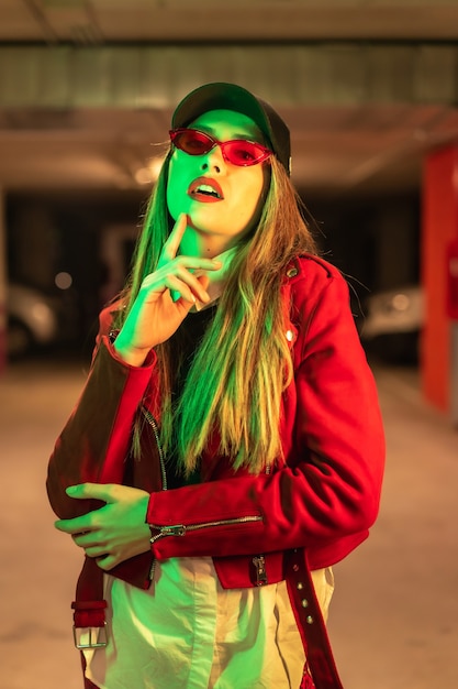 Fotografia com neons vermelhos e verdes em um estacionamento. retrato de uma jovem loira e bonita, branca, de terno vermelho e óculos escuros.