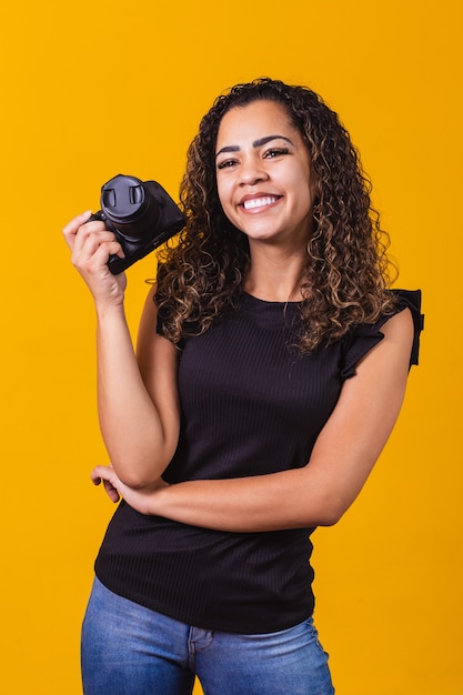 Fotografia de jovem mulher afro em fundo amarelo, segurando uma câmera fotográfica. vertical