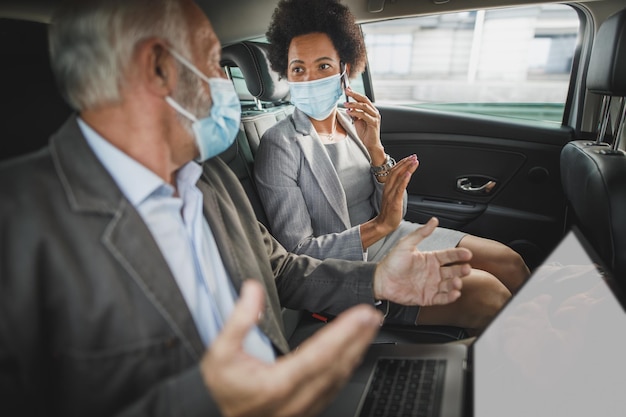 Fotografía de dos exitosos empresarios multiétnicos con máscara protectora discutiendo mientras estaban sentados en el asiento trasero de un camión en su viaje matutino durante la pandemia de COVID-19.