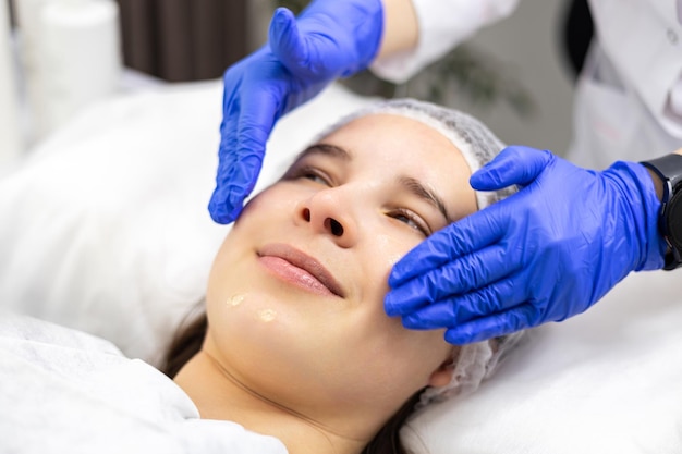Fotografia em close de uma jovem sorrindo enquanto recebe uma massagem facial de uma cosmética usando luvas de borracha Tratamento de beleza Cosmetologia estética Cuidados faciais