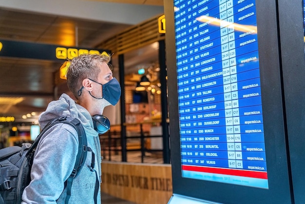 Fotografía de encabezado de un joven con mascarilla con maleta comprobando el estado de cancelación del vuelo en el tablero de información del aeropuerto en el aeropuerto vacío. Aerolínea en quiebra, crisis de la aerolínea o nuevo concepto normal