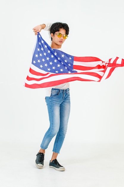 Fotografía de estudio de un joven asiático LGBT gay bisexual homosexual masculino modelo de moda con ropa informal y gafas de sol de moda de pie sosteniendo ondeando la bandera nacional de EE. UU. Sobre fondo blanco