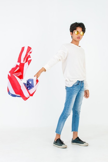 Fotografía de estudio de un joven asiático LGBT gay bisexual homosexual masculino modelo de moda con ropa informal y gafas de sol de moda de pie sosteniendo ondeando la bandera nacional de EE. UU. Sobre fondo blanco