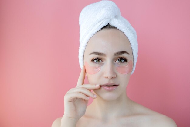 Foto fotografía de estudio de una mujer caucásica satisfecha con pecas que lleva una toalla blanca en la cabeza con parches de colágeno debajo de los ojos de pie desnuda contra un fondo rosa concepto de producto cosmético para el cuidado de la piel