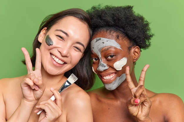 Fotografía en interiores de mujeres que muestran el signo de la paz tienen una piel suave e impecable, muestran el signo de la paz, aplican una mascarilla de arcilla nutritiva en la cara aislada sobre una pared verde