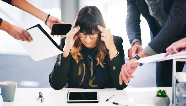 Fotografía de una joven empresaria que parece estresada mientras se siente abrumada por las solicitudes de sus colegas en una oficina moderna