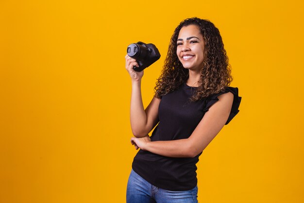 Fotografía de joven mujer afro sobre fondo amarillo sosteniendo una cámara de fotos.