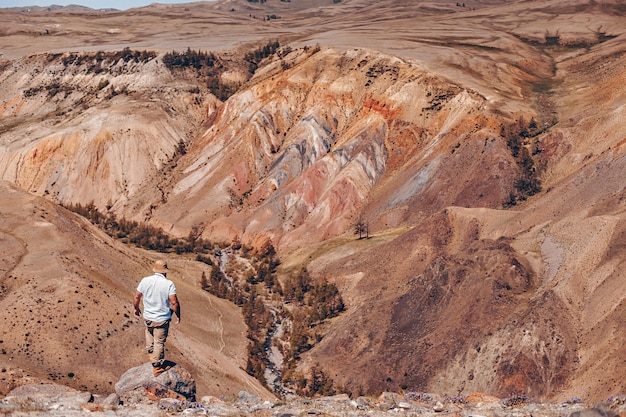 Una fotografía lejana de un hombre adulto de pie con la espalda apoyada en un adoquín al borde de un acantilado mirando una hermosa vista del cañón.