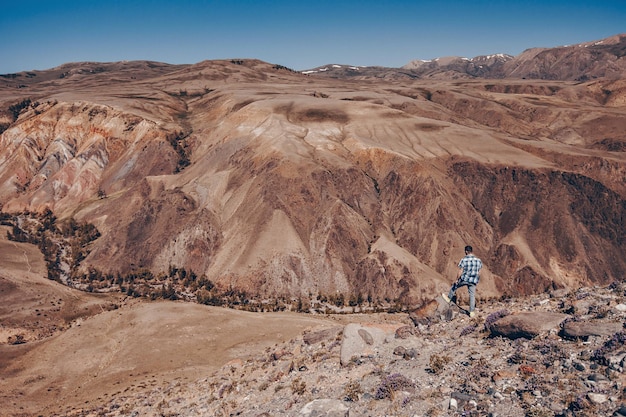 Una fotografía lejana de un hombre joven con una camisa a cuadros de pie con la espalda apoyada en un adoquín mirando el vasto paisaje de las Montañas Rocosas.