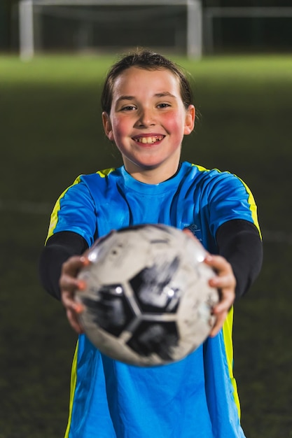 Fotografia média de uma adolescente alegre segurando uma bola de futebol em direção a uma câmera no treino da noite.