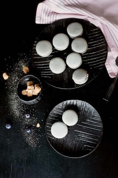 Fotografia monocromática escura Macaroons de bolos cinza na superfície escura da mesa ao lado de uma colher e açucareiro com açúcar mascavoE toalhas rosa de pano simples