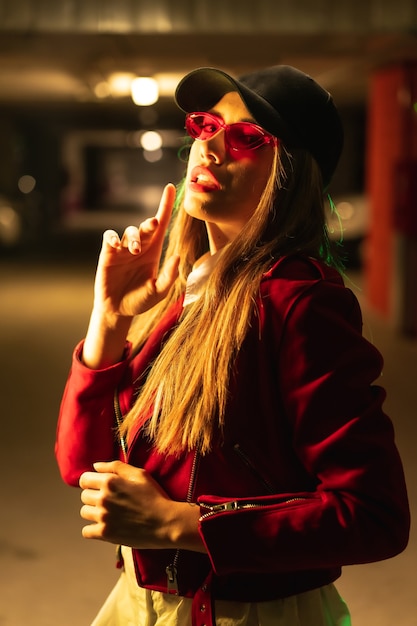 Fotografía con neones rojos y verdes en un estacionamiento. Joven mujer caucásica bastante rubia con un traje rojo, gafas de sol y una gorra negra