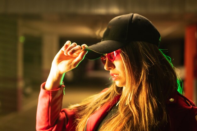 Fotografía con neones rojos y verdes en un estacionamiento. Retrato de una joven mujer caucásica rubia bonita con un traje rojo, gafas de sol y una gorra negra