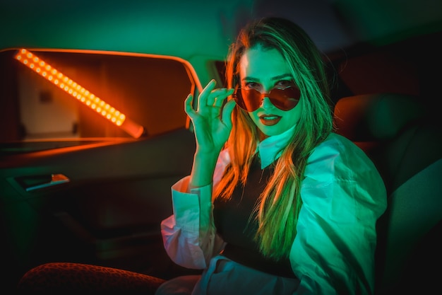 Fotografía con neones rojos y verdes en la parte trasera de un coche de una joven rubia caucásica con gafas de corazón