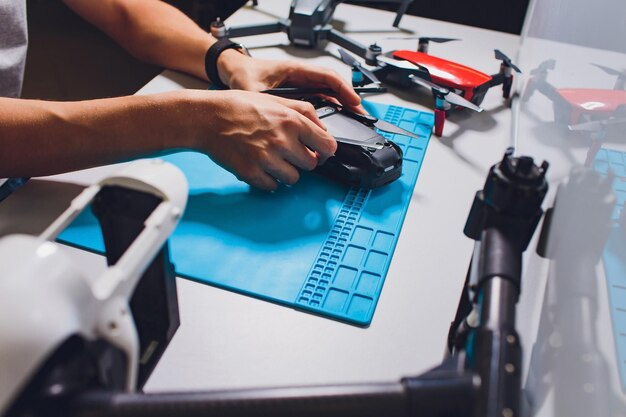 Fotografía en primer plano de un hombre trabajando en el ensamblaje de un nuevo sistema de vigilancia utilizando un dron quadcopter con cámara en la mesa con diferentes herramientas en un taller moderno