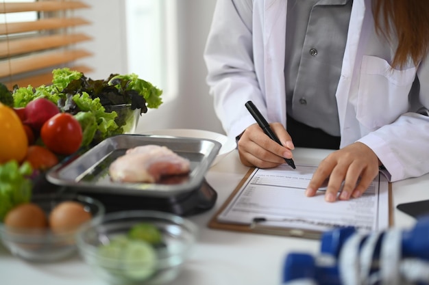 Fotografía recortada de una dietista sentada en un escritorio con diferentes productos saludables y escribiendo un plan de dieta