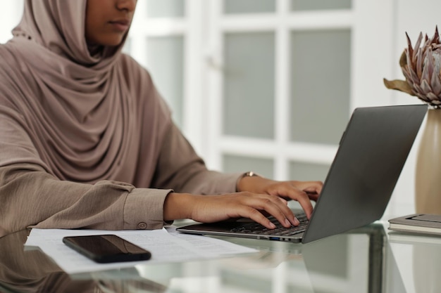 Fotografía recortada de una joven mujer de negocios con hijab escribiendo en el teclado de una computadora portátil