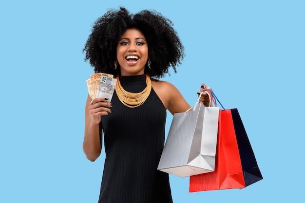 Fotografía de una sonriente joven mujer afro sosteniendo bolsas de la compra sobre fondo azul.