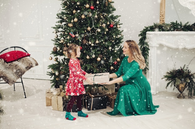 Fotografía de Stock de amorosa madre en vestido verde dando a su pequeña hija en pijama vestido un regalo de Navidad. Están al lado de un árbol de Navidad bellamente decorado bajo la nieve.