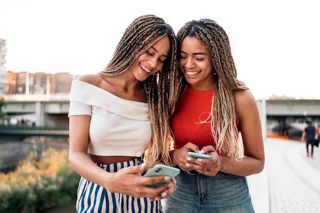 Fotografía de Stock de jóvenes hermanas afroamericanas riendo y usando sus teléfonos en la calle