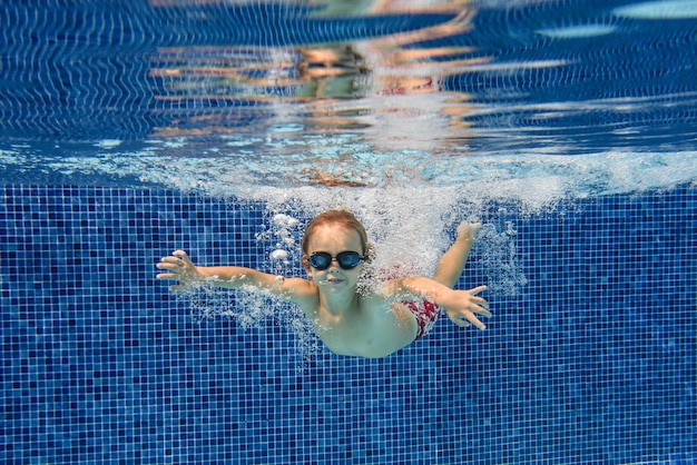 Fotografía submarina de un niño pequeño con gafas nadando en agua limpia y transparente de la piscina y mirando la cámara contra el fondo azul