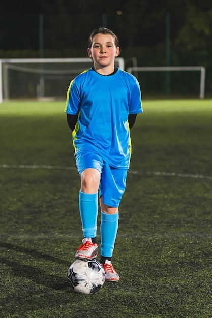 Fotografia vertical completa de uma bonita jogadora de futebol caucasiana em uniforme azul posando com uma bola