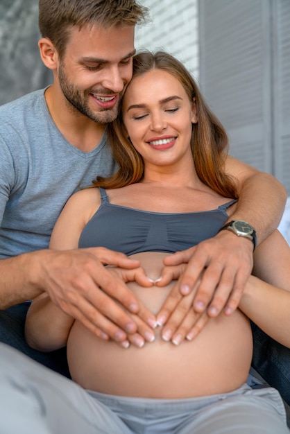 Fotografia vertical de um jovem casal fazendo uma forma de coração na barriga grávida com as mãos