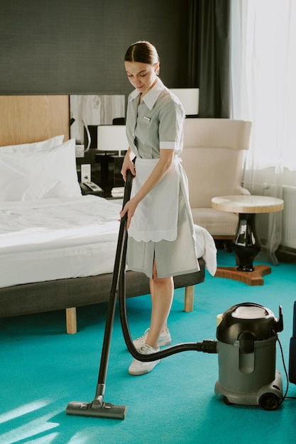 Fotografia vertical de uma jovem camareira profissional usando um aspirador moderno para limpar o chão de um hotel