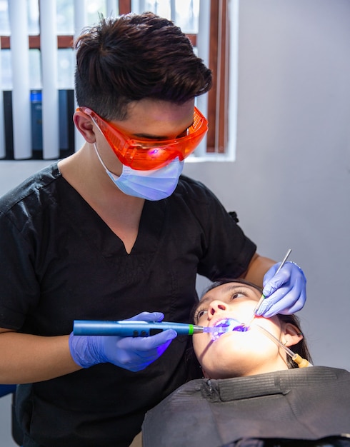 Fotografía vertical de un paciente en el consultorio de un dentista que recibe un procedimiento de blanqueamiento dental que tiene PPE con máscara, guantes y gafas de color naranja