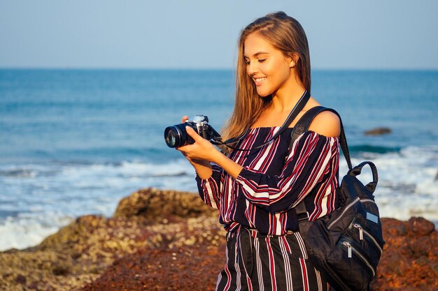 Fotografía y viajes Mujer joven en elegante mono a rayas sosteniendo cámara hermosa vista tropical del mar y las rocas chica europea atractiva con un hombro de mochila fotografiando el paisaje del paraíso